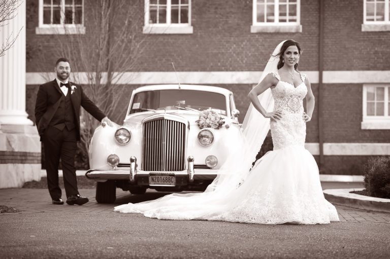 Bride in a white wedding gown standing next to a groom in a black tuxedo beside a classic white limousine.
