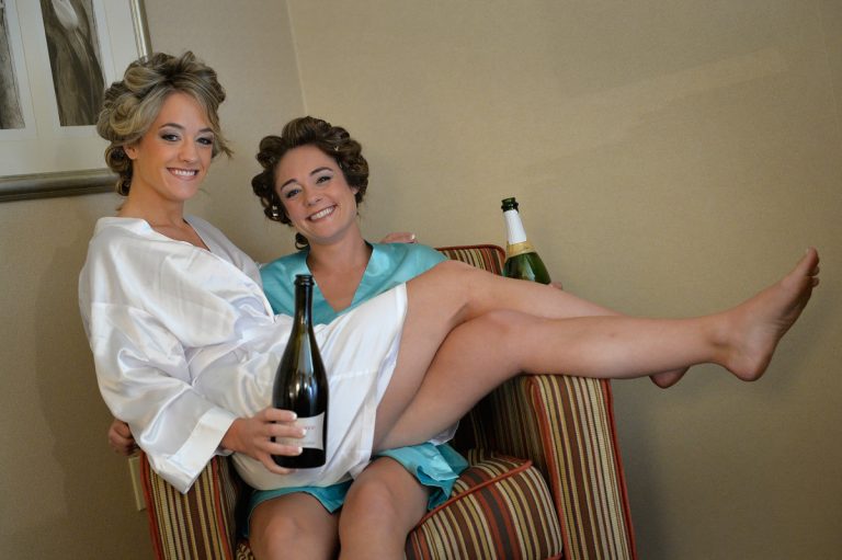 Two women sitting on a chair, wearing robes and holding champagne bottles, smiling at the camera.