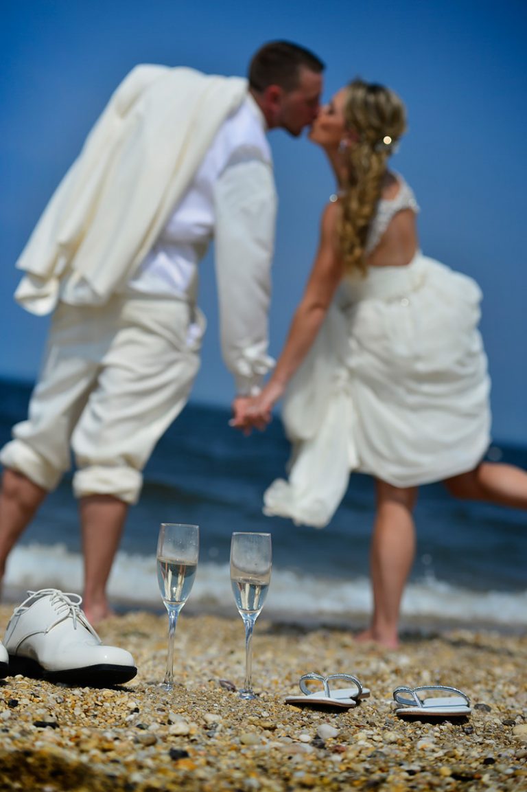 A couple in wedding attire kissing on the beach with champagne glasses in the foreground.