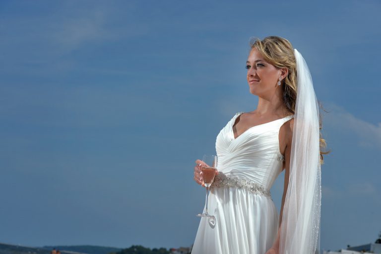 Bride wearing a white wedding dress and veil, holding a glass, against a blue sky.