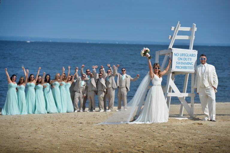 Bridal party posing on the beach with a lifeguard stand displaying a swimming prohibition sign.