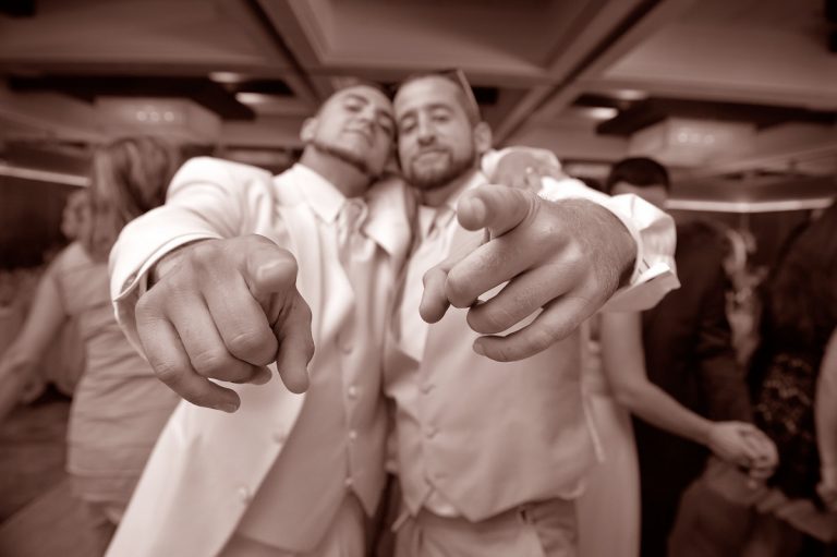 Two men in formal attire pointing at the camera with smiles during a celebration.