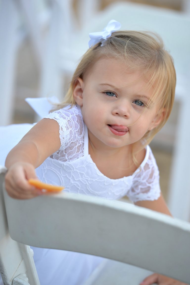 Young girl wearing a white dress and a bow in her hair, holding a snack while sitting on a chair.