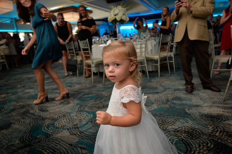 A young girl wearing a white dress with a bow, standing in a crowded event space.