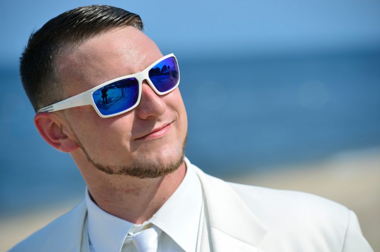 Groom wearing a white suit and sunglasses with a beach background.