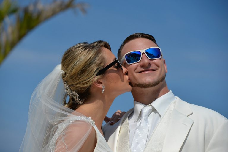 Bride and groom wearing sunglasses, with the bride kissing the groom on the cheek.