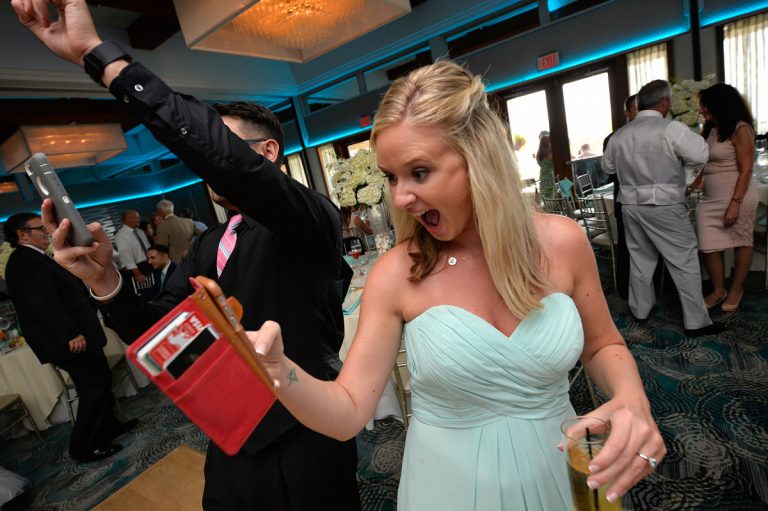 A woman in a light blue dress excitedly holds a red book while a man takes a photo at a wedding reception.
