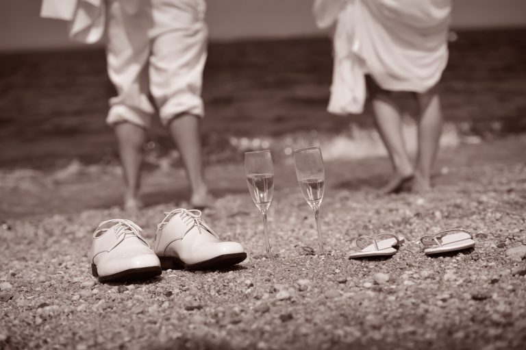 White wedding shoes and champagne glasses on a sandy beach with ocean in the background.