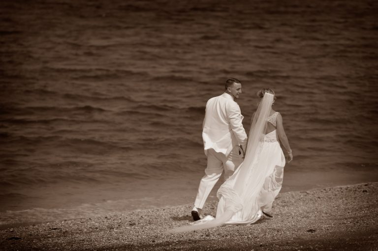 A couple in wedding attire walking along a beach with water in the background.