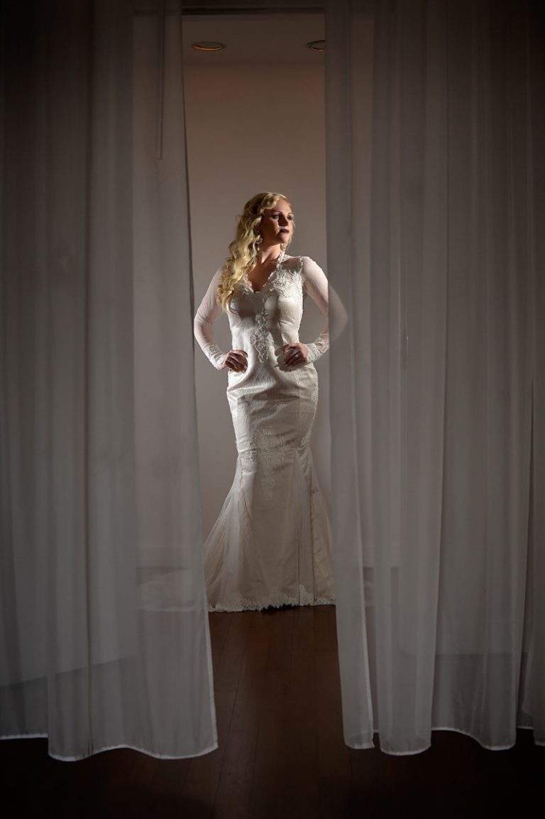 Bride wearing an elegant white wedding dress with long sleeves, standing behind sheer curtains.