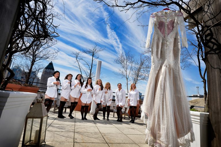 Group of bridesmaids in white robes standing on a rooftop with a wedding dress hanging nearby.