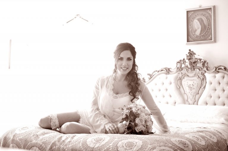 A bride in a white outfit sitting on a bed with a bouquet of flowers in her hand.