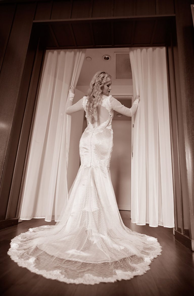 A bride wearing a fitted lace wedding dress with a long train, standing with her back to the camera.