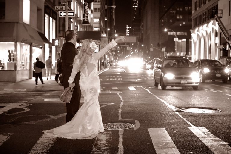 Bride in a wedding dress hailing a taxi on a busy New York City street at night.