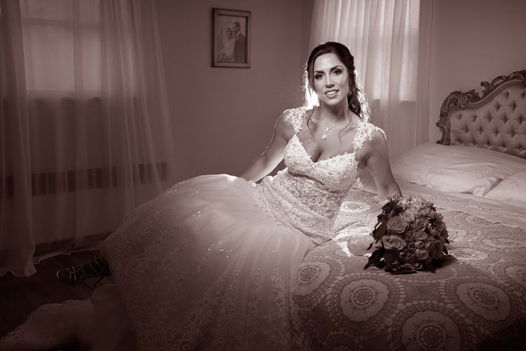 Bride wearing a wedding dress, sitting on a bed with a bouquet of roses.