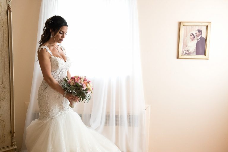 Bride in a wedding dress holding a bouquet of flowers, standing by a window with a portrait in the background.