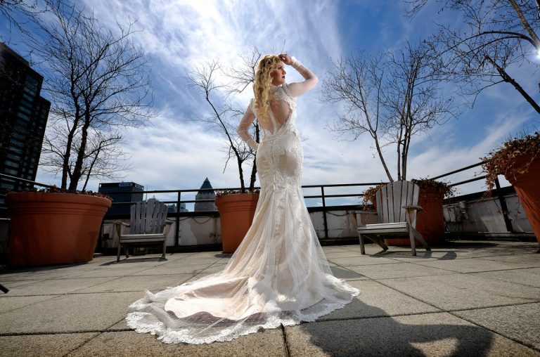 Bride wearing an elegant white dress with a long train, posing on a rooftop with a city skyline in the background.