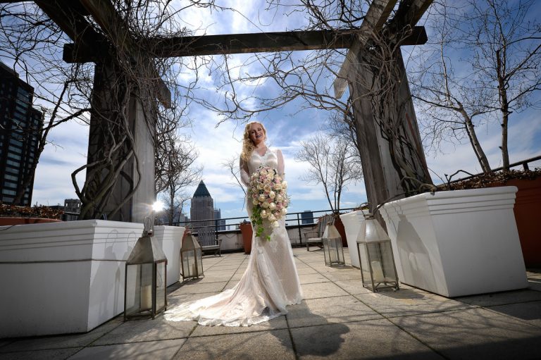 Bride in a white gown holding a bouquet of flowers on a rooftop with a city skyline in the background.