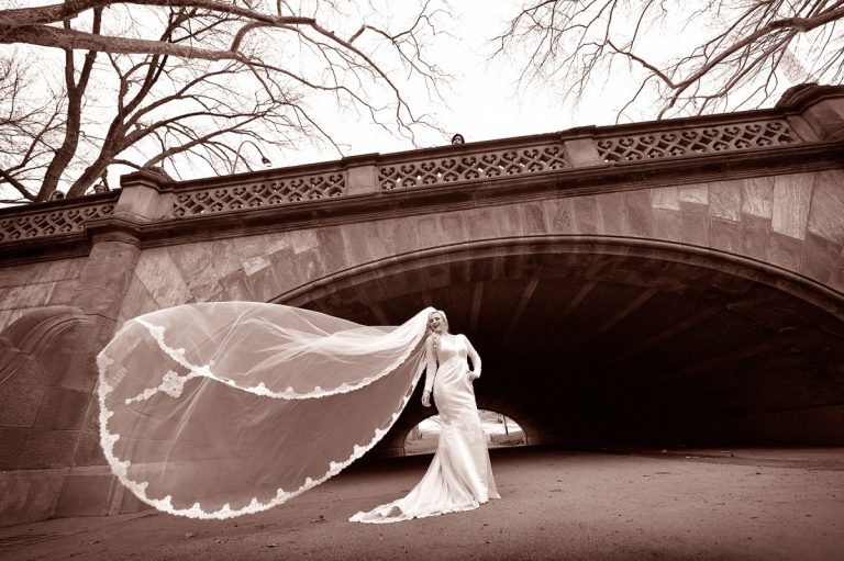 Bride wearing a white wedding dress and veil, standing under a stone bridge in Central Park.