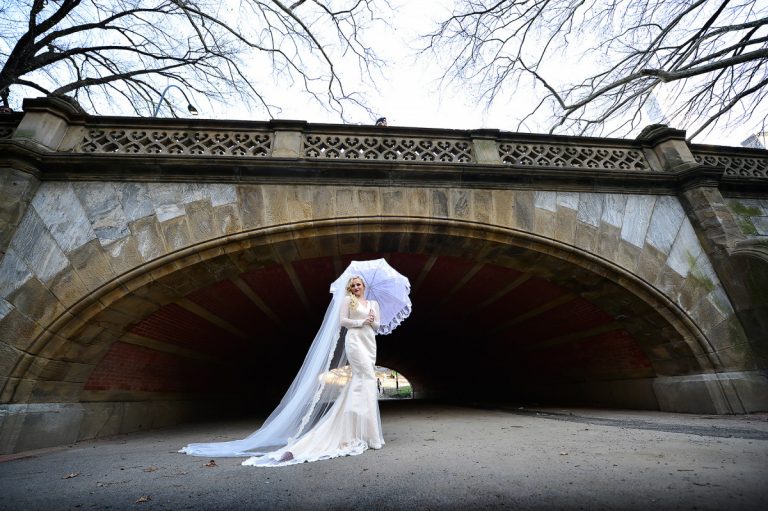 Bride holding a white umbrella while standing under a stone arch in Central Park, NYC.