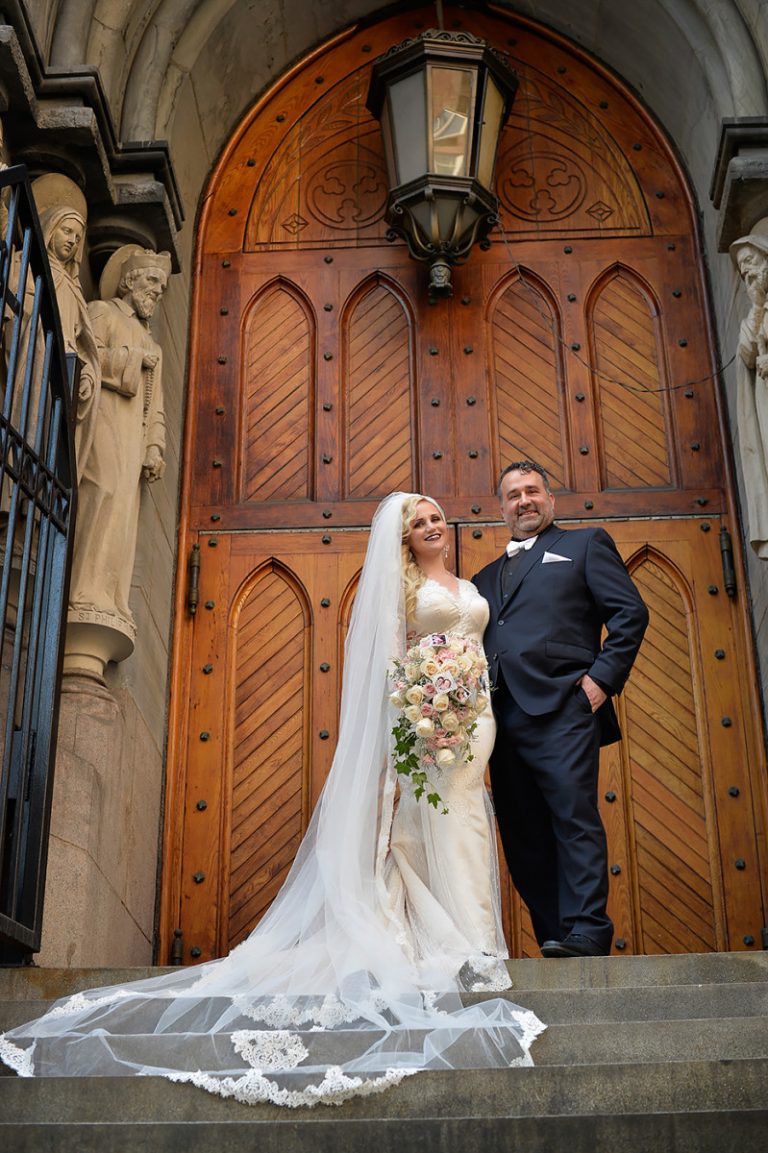 Bride in a wedding dress and groom in a tuxedo standing at a church entrance with wooden doors.