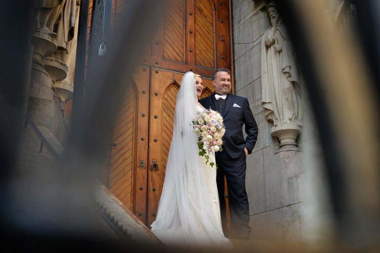 Bride in a white gown and groom in a black tuxedo standing at a church entrance with a floral bouquet.