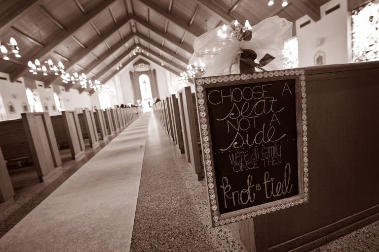 Interior of a church featuring wooden pews and a decorative sign that reads 'Choose a seat, not a side, we're all family once the knot is tied.'