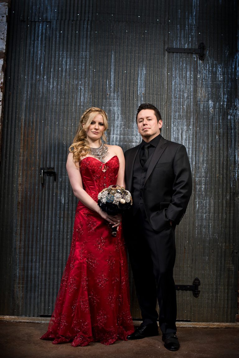 Bride wearing a red dress and groom in a black suit standing together in front of a textured background.