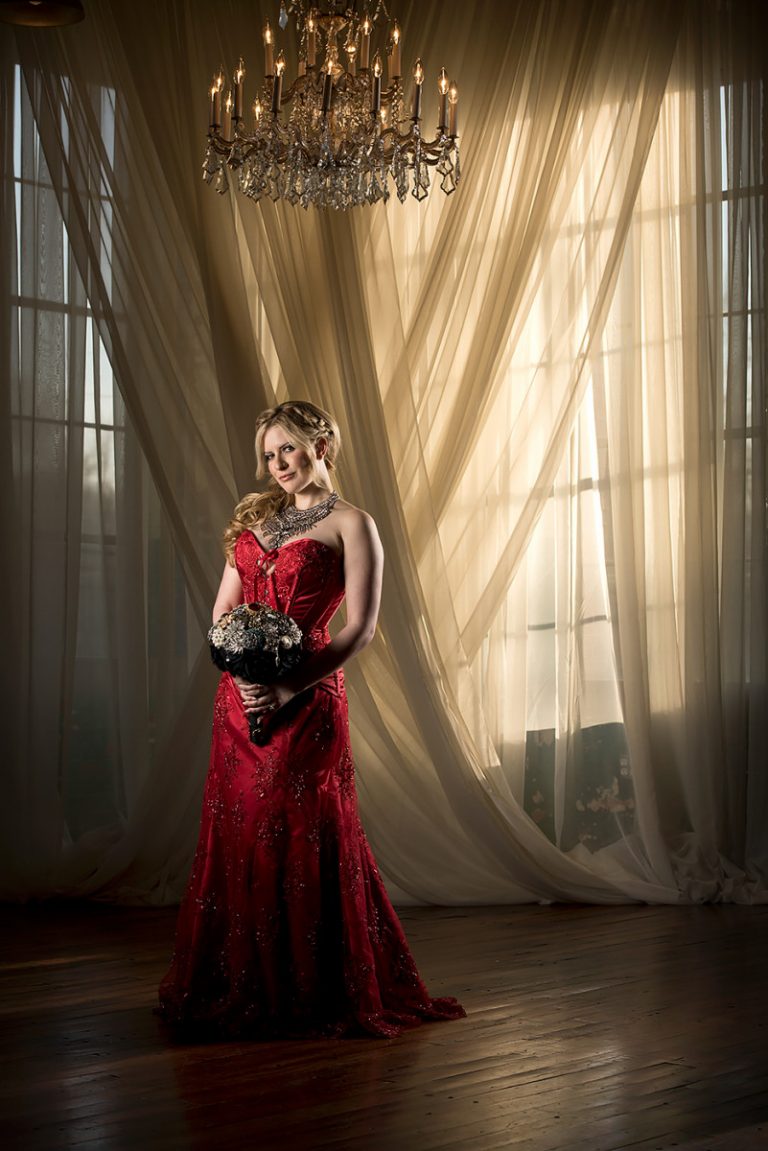 Bride wearing a red Gothic Victorian wedding dress, holding a bouquet, with a chandelier above.