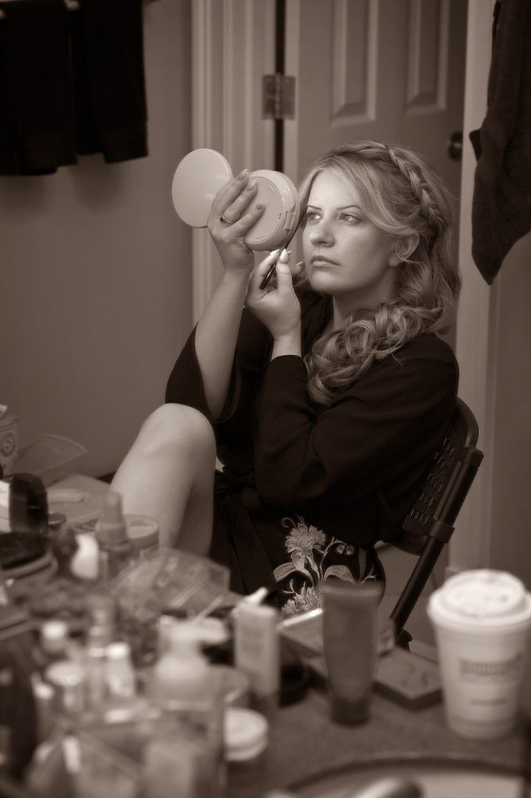 A woman with long hair styled in waves looking into a handheld mirror while applying makeup.
