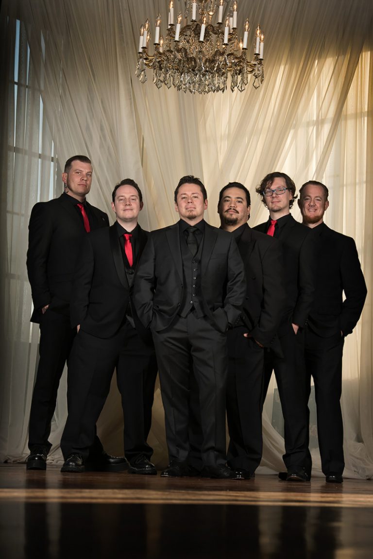 Group of six groomsmen dressed in black suits and red ties standing together under a chandelier.