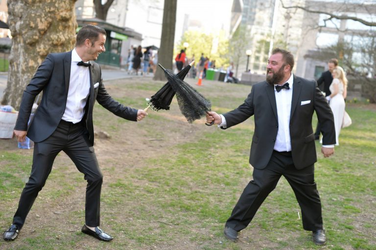 Two groomsmen in tuxedos playfully engaging in an umbrella sword fight outdoors.