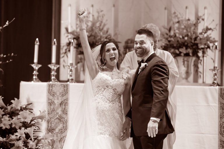Bride and groom celebrating joyfully during their wedding ceremony with raised hands.