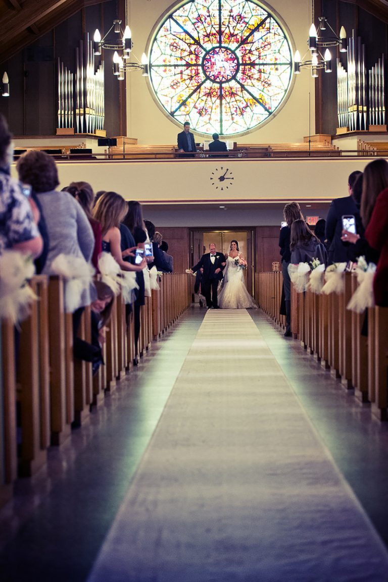 A bride walking down the aisle in a church with a stained glass window in the background.