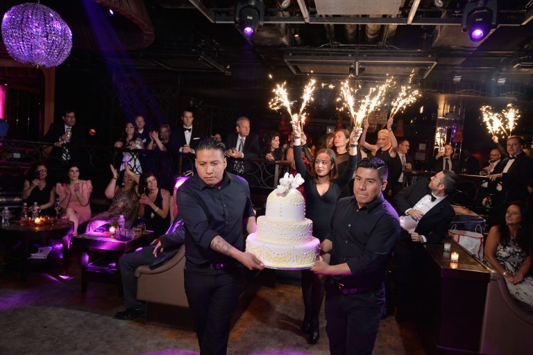 Two individuals carrying a large wedding cake adorned with flowers at Lavo in New York City.
