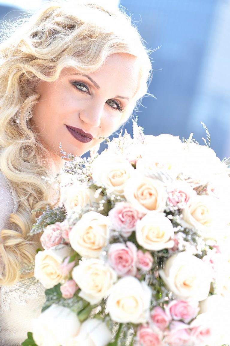 Bride with long blonde hair holding a bouquet of pink and white roses outdoors.