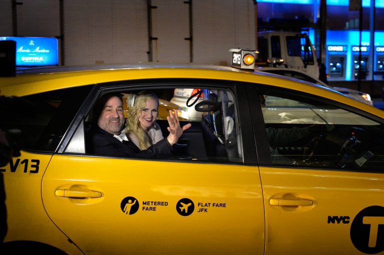Bride and groom sitting in a yellow taxi in New York City, waving at the camera.