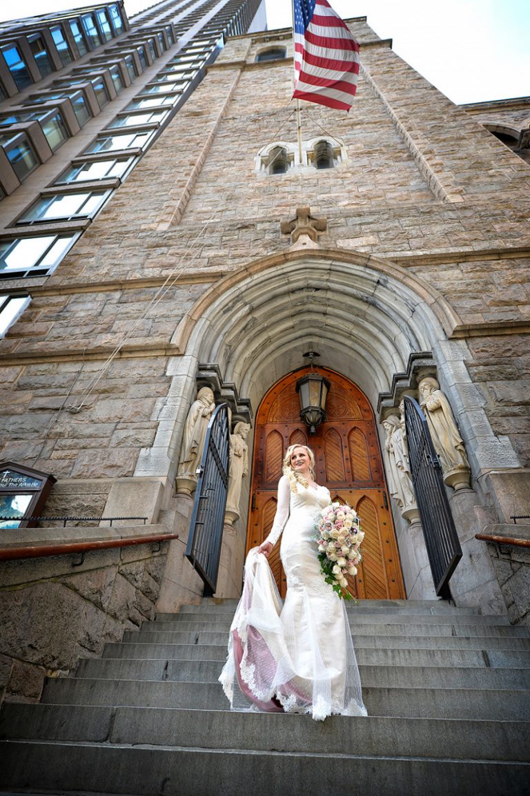 Bride in a wedding dress holding a bouquet standing at the entrance of a church with an American flag above.