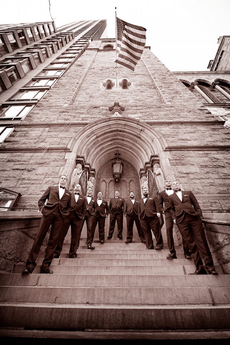 Groomsmen in formal attire standing on the steps of a church in New York City with an American flag above.