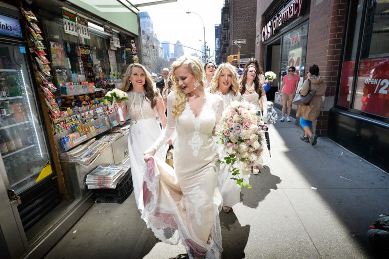 Group of bridesmaids in white dresses walking down a street in New York City, carrying bouquets.