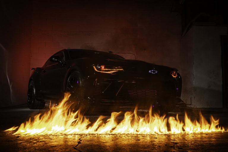 Black Chevrolet Camaro positioned in front of flames on a concrete surface, with dramatic lighting effects.