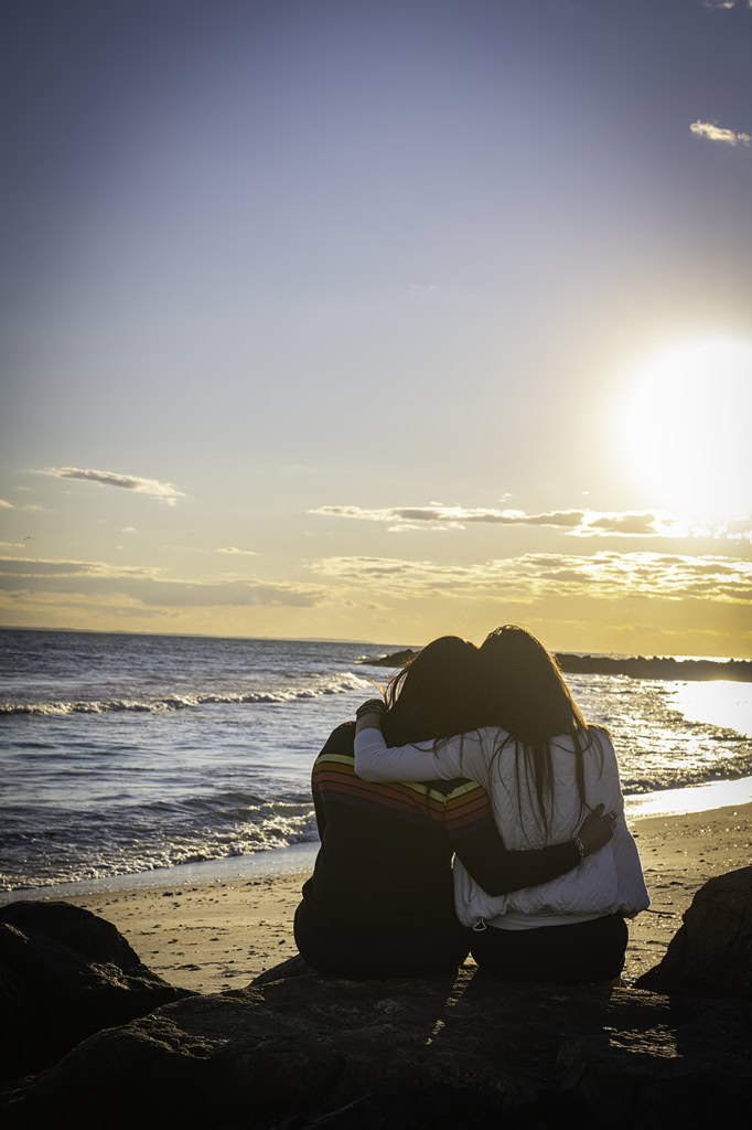 Two individuals sitting in Rockaway Beach, embracing each other while watching the sunset over the ocean.