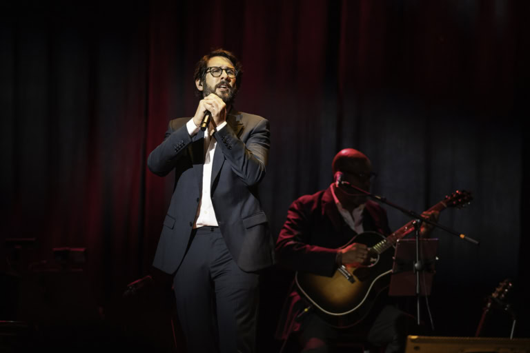 Josh Groban in a suit performing at Tilles Center, accompanied by a guitarist in the background.