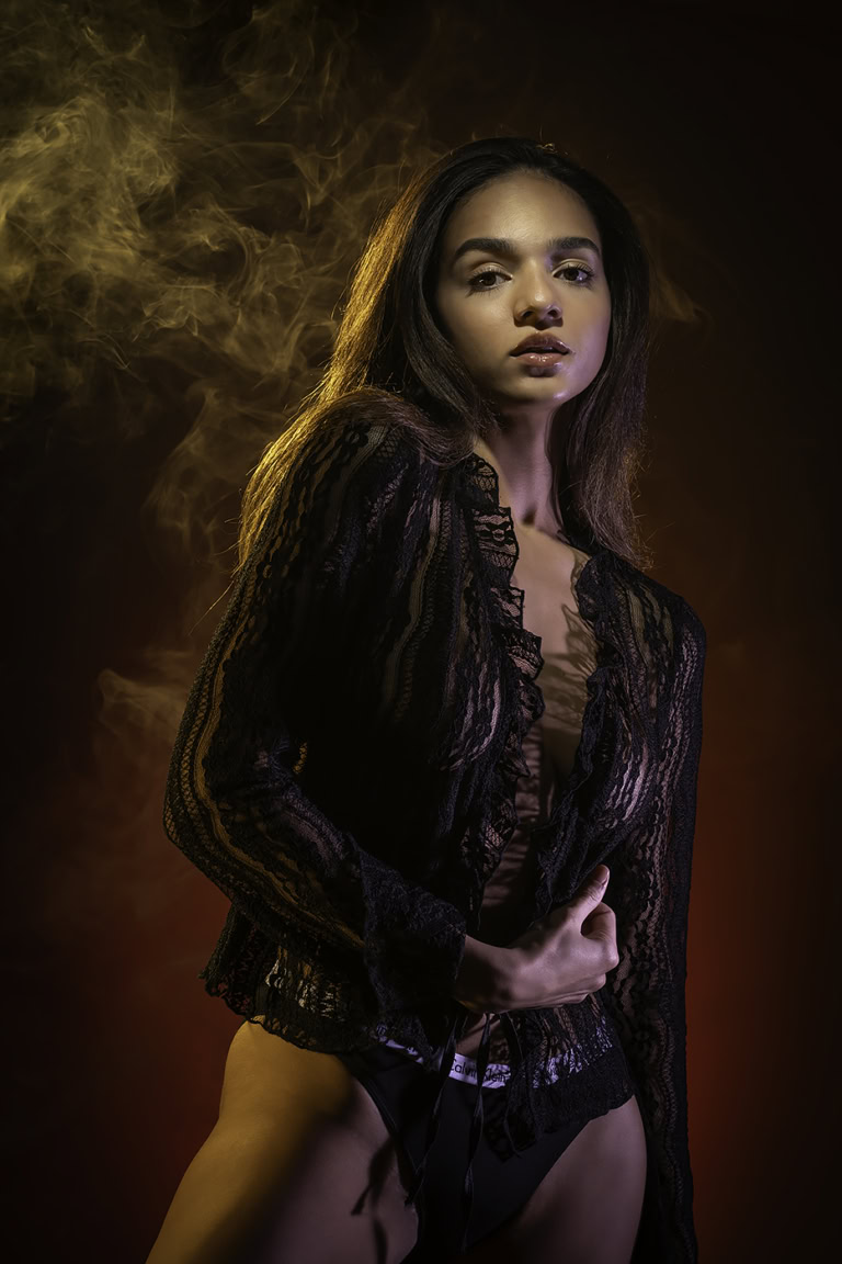 Model posing in a black lace garment with a smoky background, showcasing intricate lace details and a confident expression.