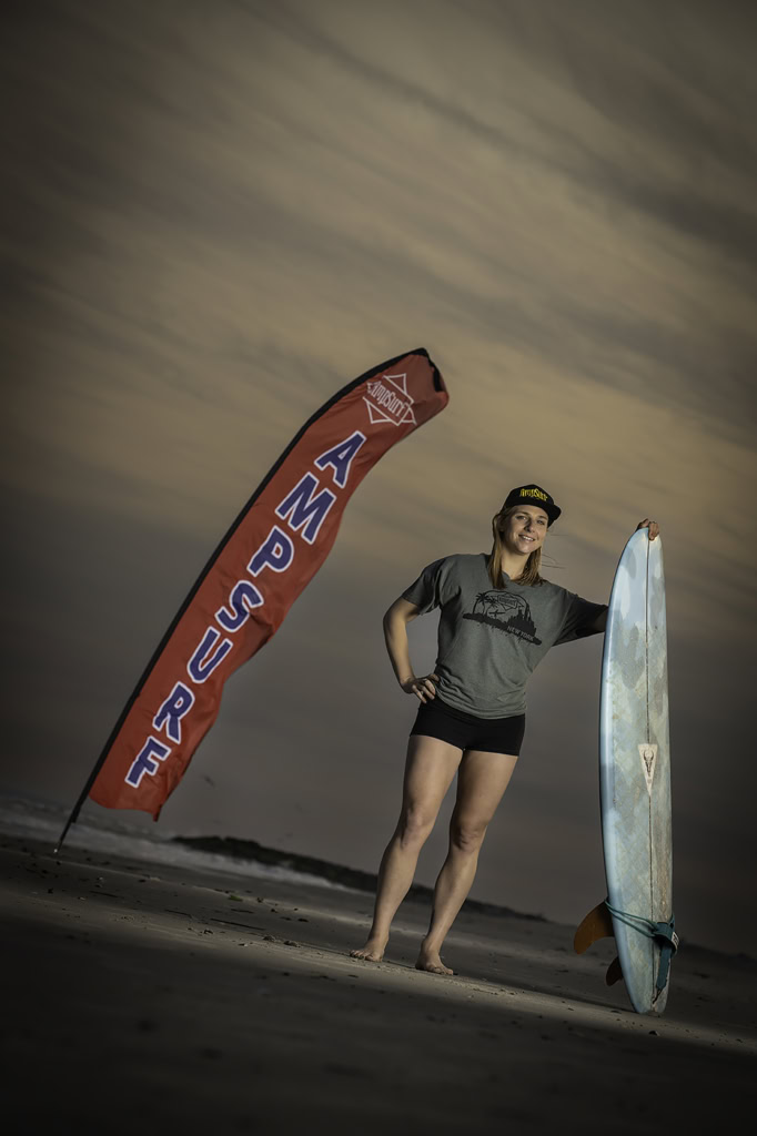 Ashley standing on a beach holding a surfboard next to a red flag with 'AMPSURE' printed on it.