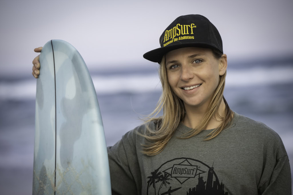 Ashley wearing a black cap with 'AmpSurf' logo and a gray shirt, holding a surfboard in Rockaway Beach