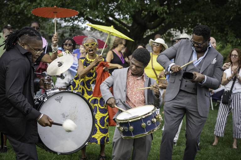 Group of musicians playing drums and celebrating outdoors with colorful umbrellas and vibrant attire.