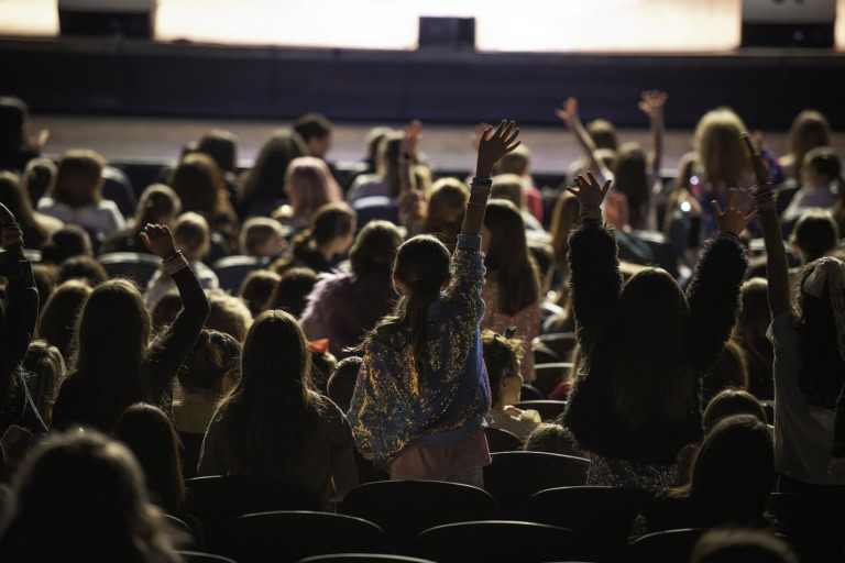 Audience of children with raised hands during a performance at Tilles Center, showcasing excitement and engagement.