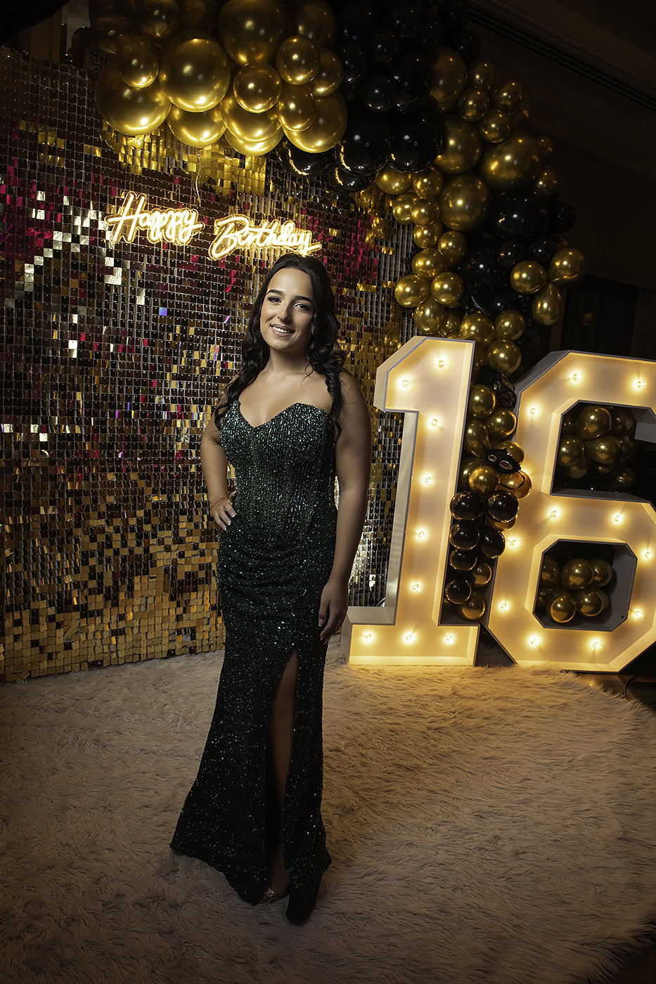 Young woman in a green sequined gown poses beside a glittering gold mosaic backdrop, with black and gold balloons and a large illuminated 18 sign at a birthday event