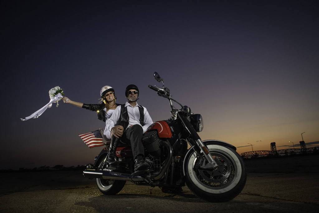 Two riders pose on a black motorcycle as an American flag waves beside them at sunset near a waterfront.